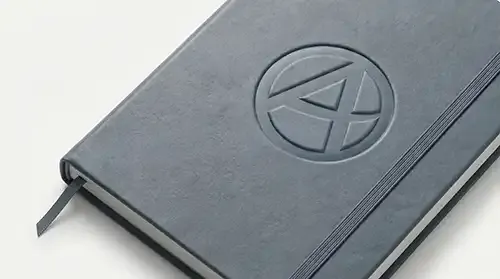 Cuaderno corporativo A5 de polipiel azul grisáceo con banda elástica vertical y logotipo corporativo abstracto en bajorrelieve en la portada 16-9
