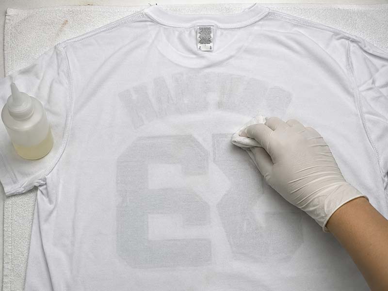 Persona con guantes quitando un estampado de una camiseta blanca usando un paño y disolvente especial