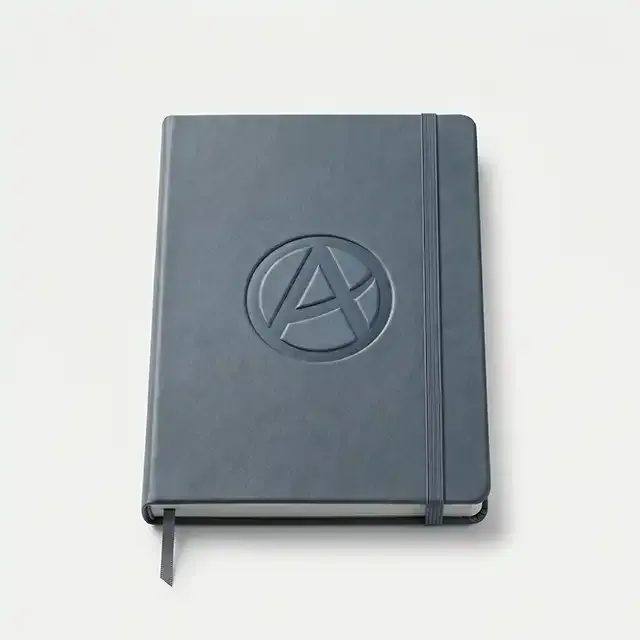 Cuaderno corporativo A5 de polipiel azul grisáceo con banda elástica vertical y logotipo corporativo abstracto en bajorrelieve en la portada.
