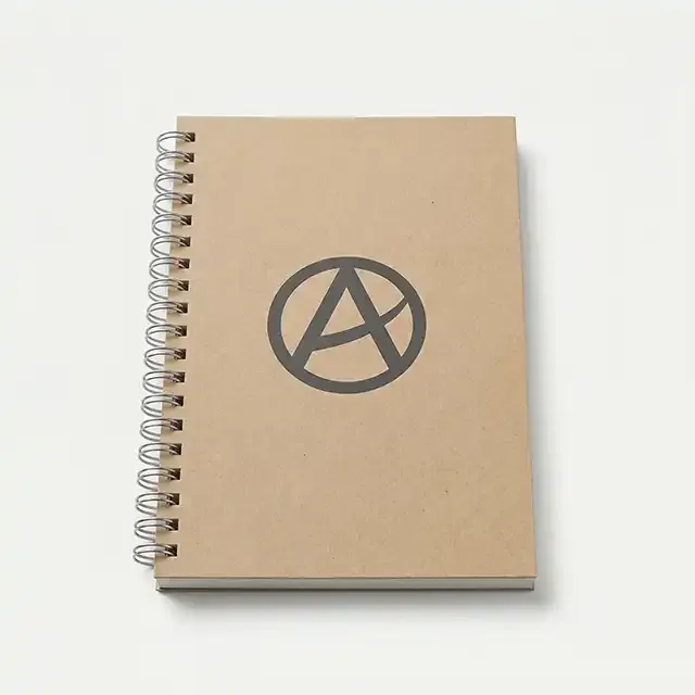 Cuaderno corporativo A5 de tapa blanda en cartón kraft natural con encuadernación de espiral metálica y logotipo serigrafiado en gris oscuro.
