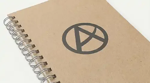 Cuaderno corporativo A5 de tapa blanda en cartón kraft natural con encuadernación de espiral metálica y logotipo serigrafiado en gris oscuro 16-9