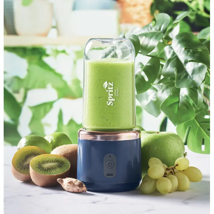 Licuadora portátil azul con batido verde rodeada de kiwis, manzana verde, uvas y jengibre en un entorno de cocina con plantas