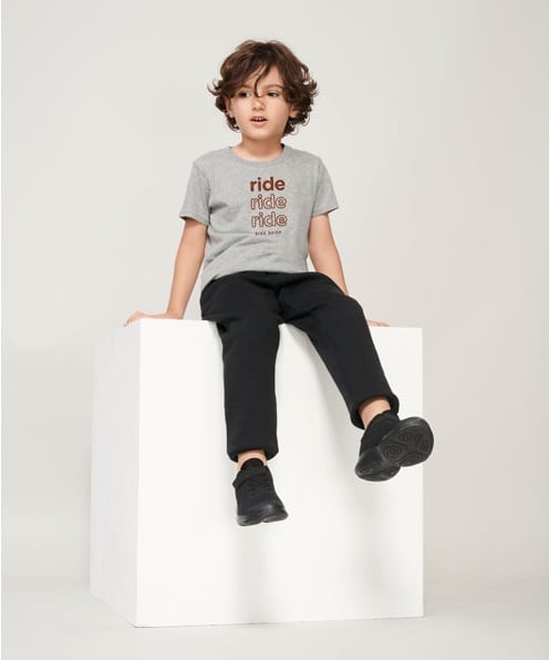 Niño sentado con camiseta gris de algodón con texto marrón y pantalones negros.