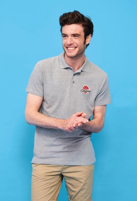 Hombre con polo gris de manga corta con logo rojo y beige posando sobre fondo azul.