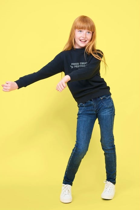 Niña pelirroja con sudadera negra con texto, vaqueros azules y zapatillas blancas sobre fondo amarillo.