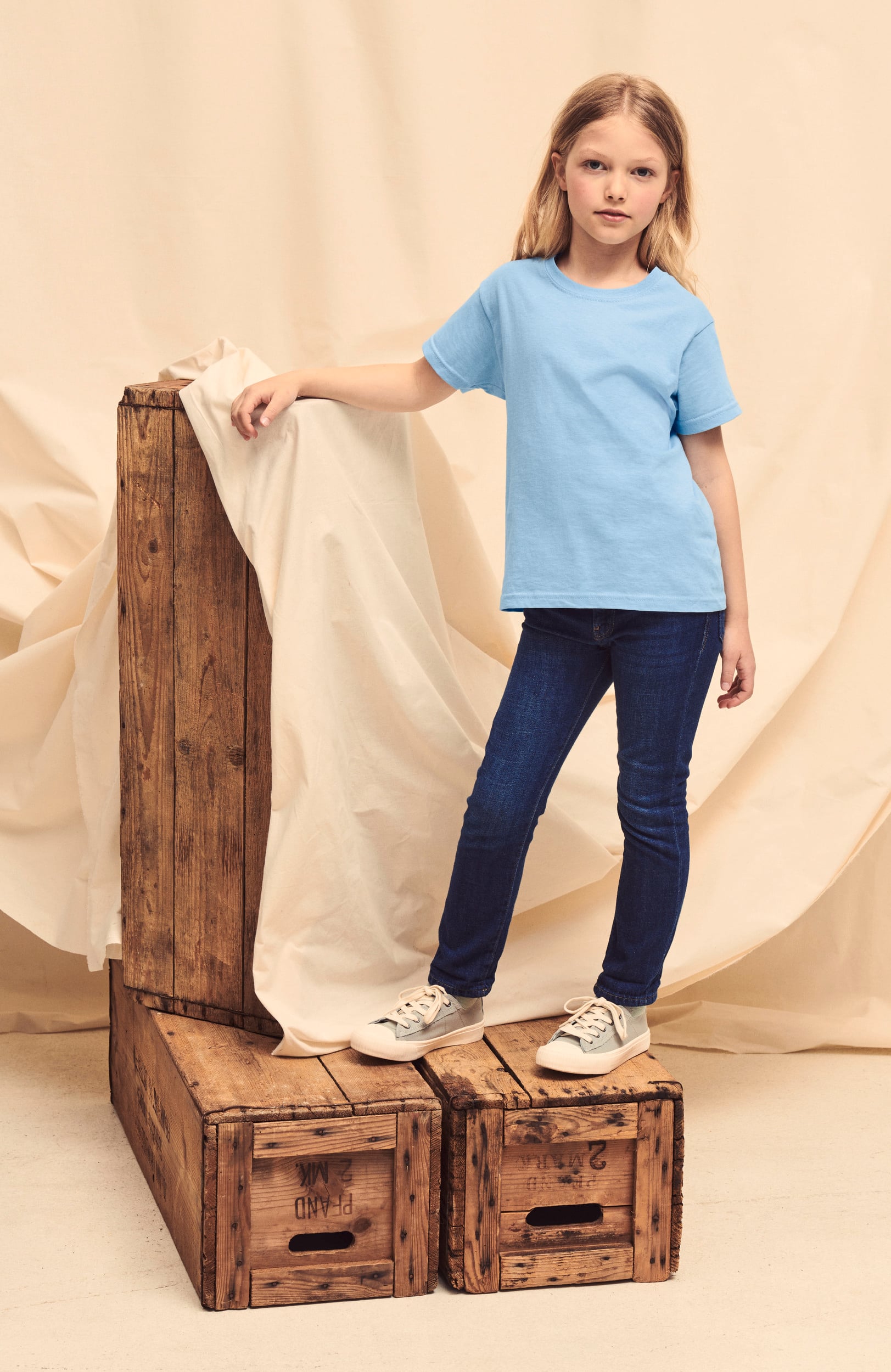 Niña con camiseta azul claro, vaqueros oscuros y zapatillas blancas de lona apoyada en cajas de madera.