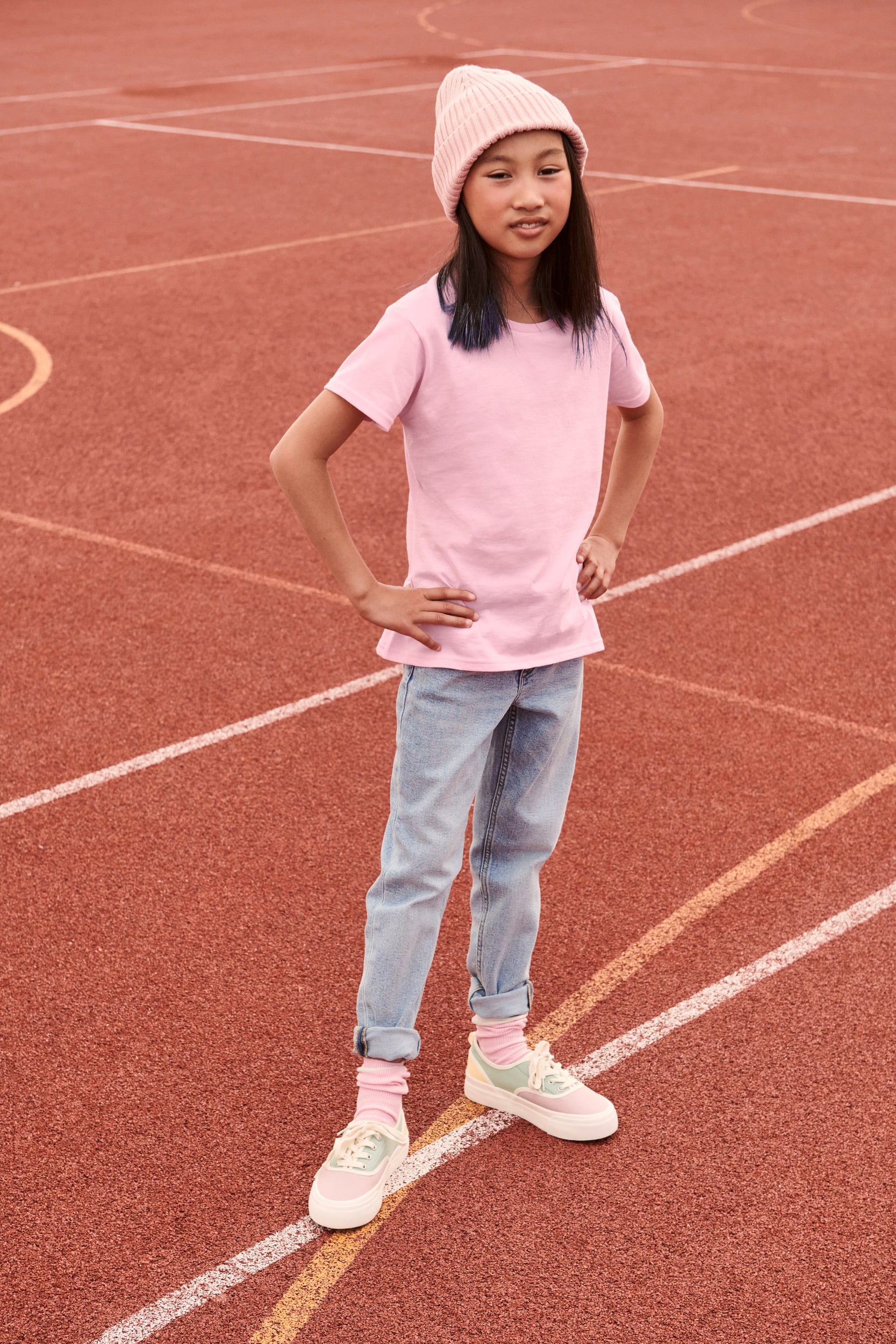Niña con camiseta rosa, vaqueros claros, calcetines y zapatillas multicolor sobre pista deportiva.
