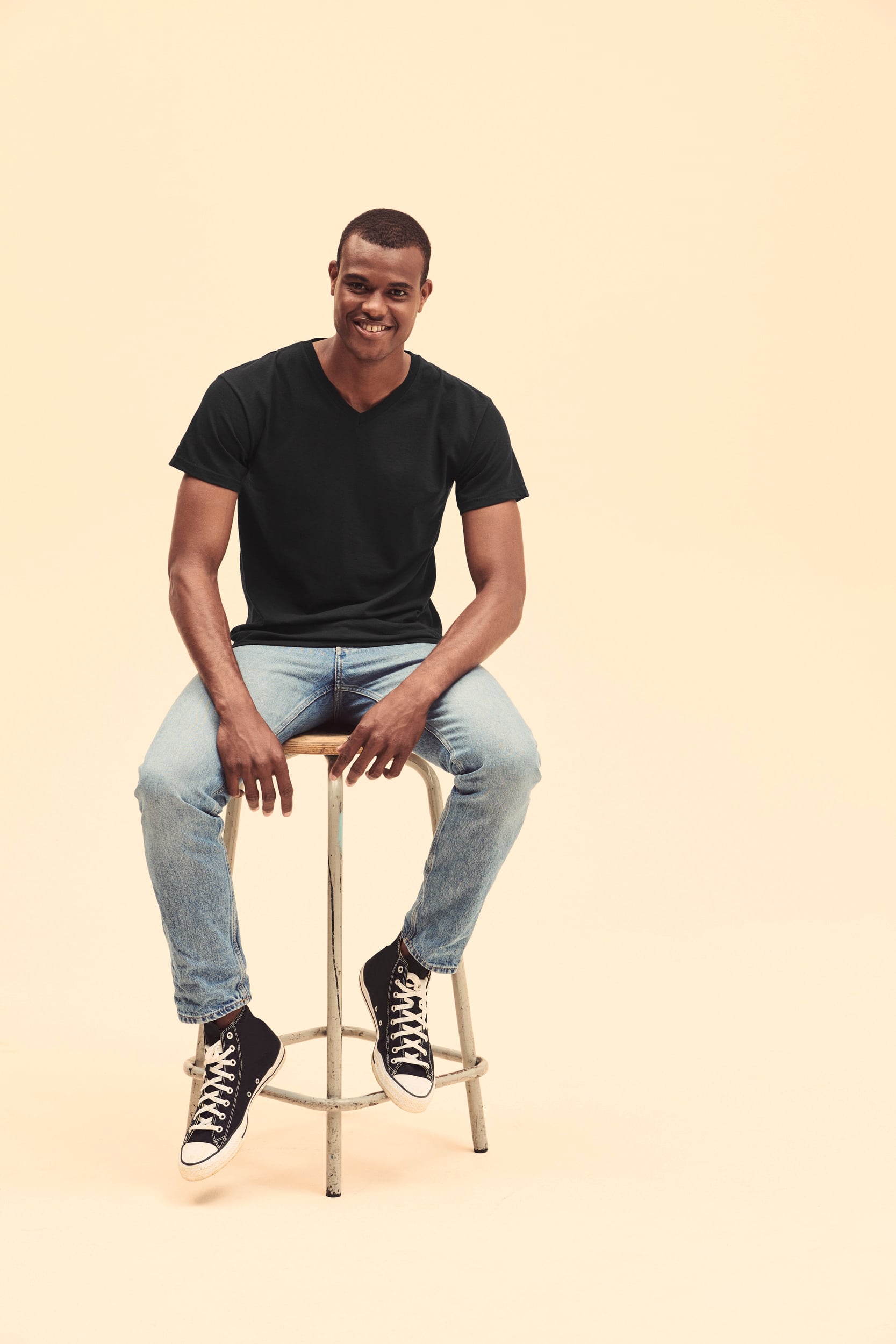Hombre sentado en taburete con camiseta negra de manga corta, vaqueros claros y zapatillas negras altas.