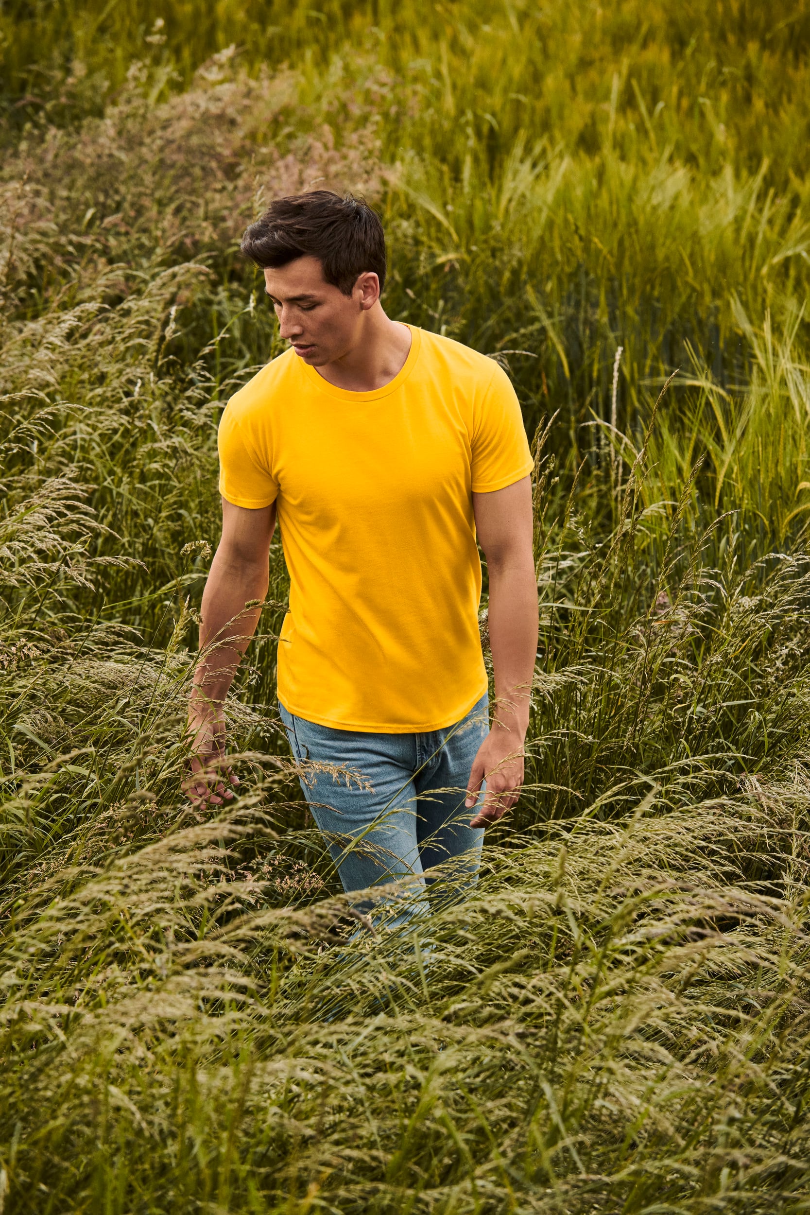 Hombre con camiseta amarilla de manga corta y vaqueros claros caminando entre hierba alta.