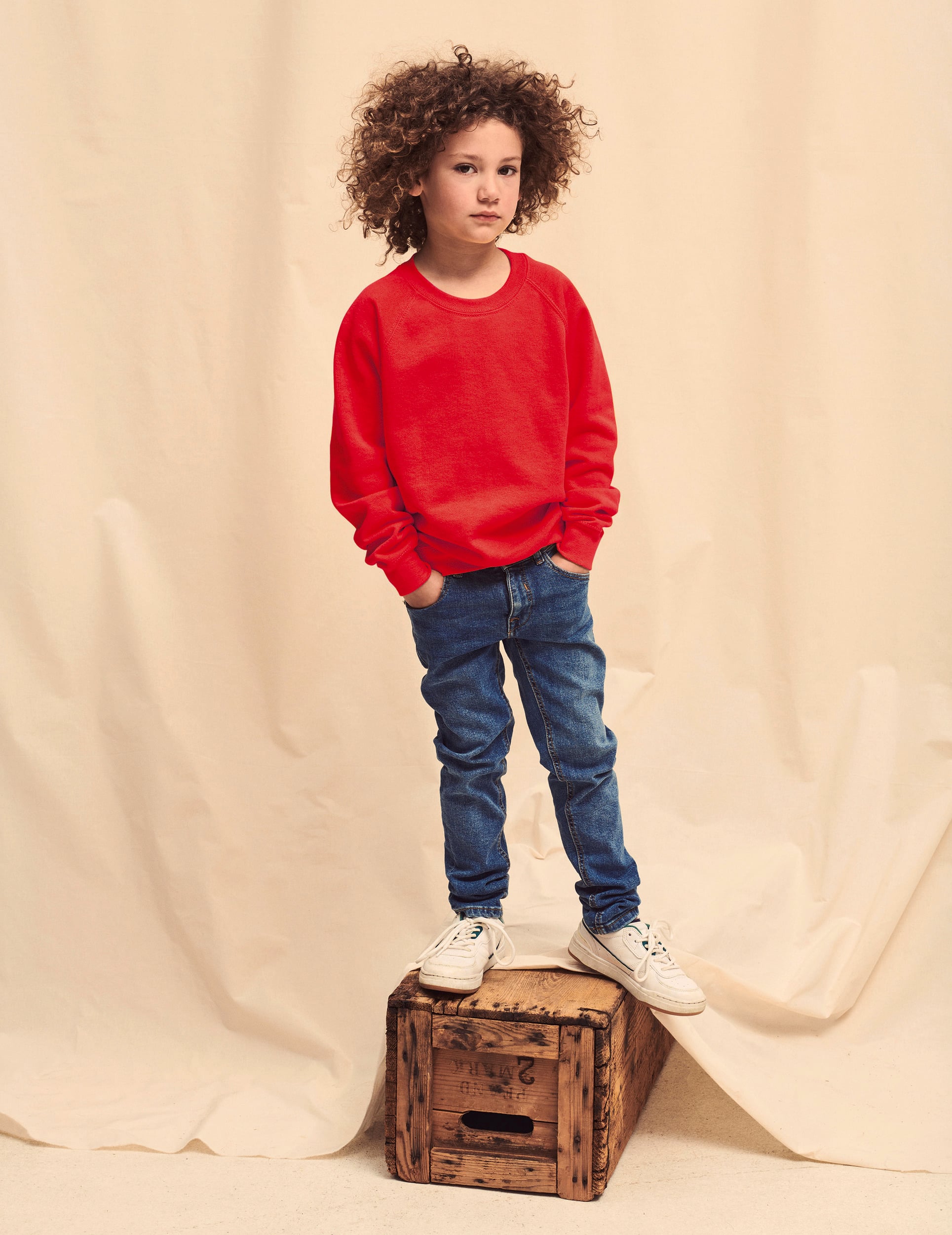 Niño con sudadera roja, vaqueros azules y zapatillas blancas de pie sobre una caja de madera.