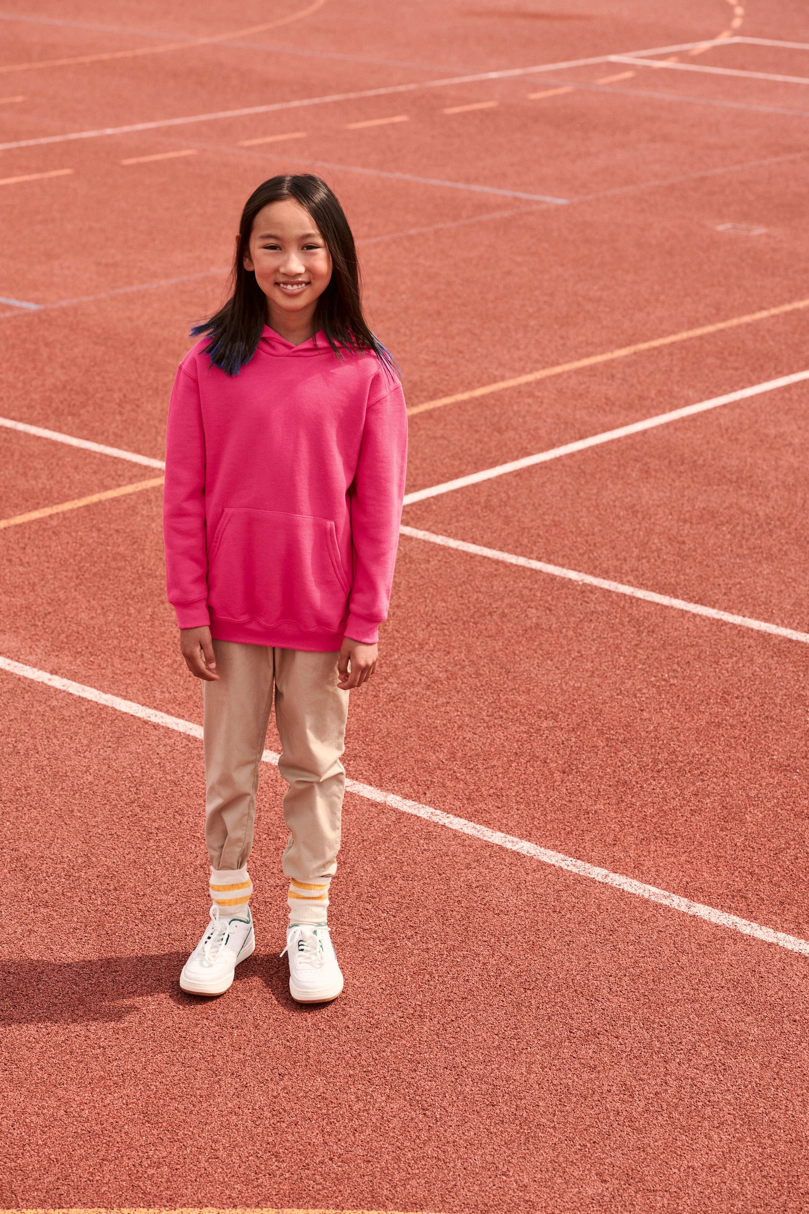 Niña con sudadera rosa, pantalones beige y zapatillas blancas en pista de atletismo.