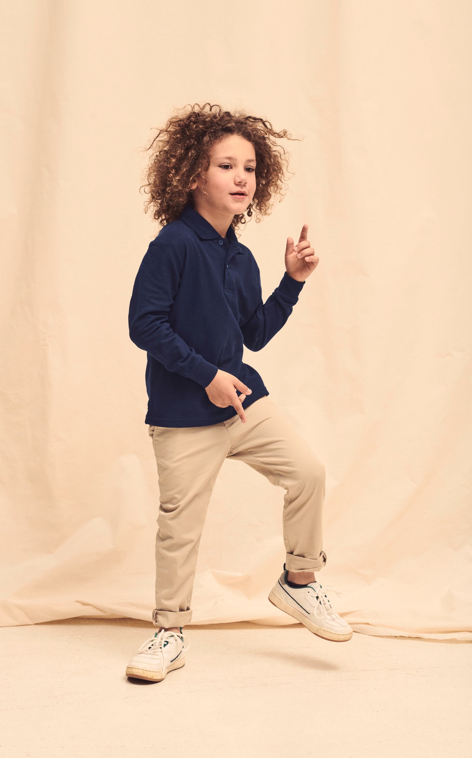 Niño con camiseta polo azul marino de manga larga, pantalones beige y zapatillas blancas deportivas.