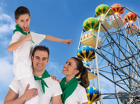 Familia con pañuelos verdes sonriente junto a una noria de globos de colores en un parque de atracciones.